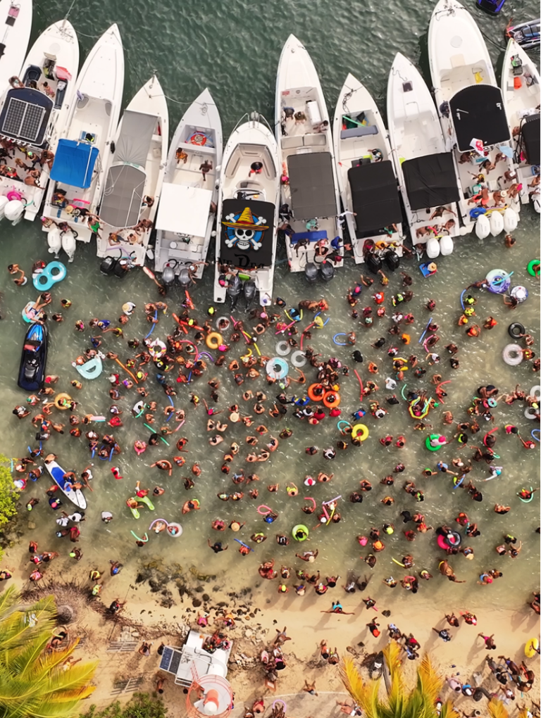 Vue aérienne d’une foule de personnes rassemblées dans l’eau et sur la plage lors du Tour des Yoles 2025 en Martinique, entourées de bateaux, bouées colorées et musique festive